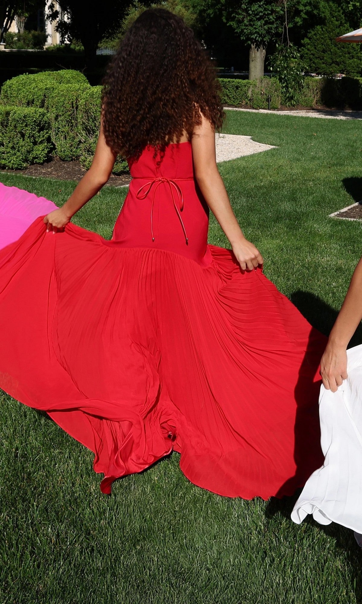 A person with curly hair stands on grass holding out the sides of their long red Jovani 47000 formal dress, which features a gold floral waist detail. Other people in pink and white dresses are partially visible on either side.